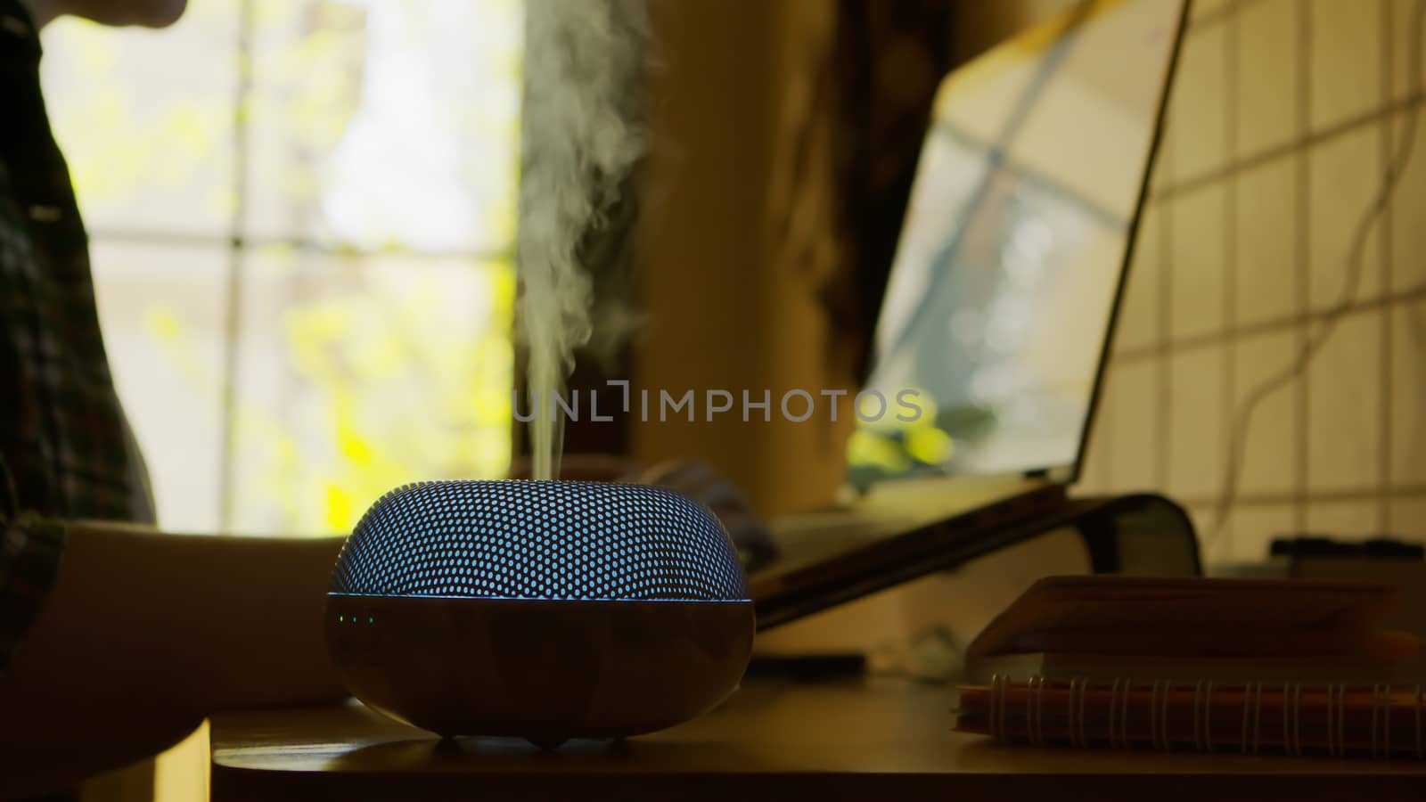 Vapor coming from blue led esential oil diffuser with aromatic perfume while freelance woman is working on laptop.
