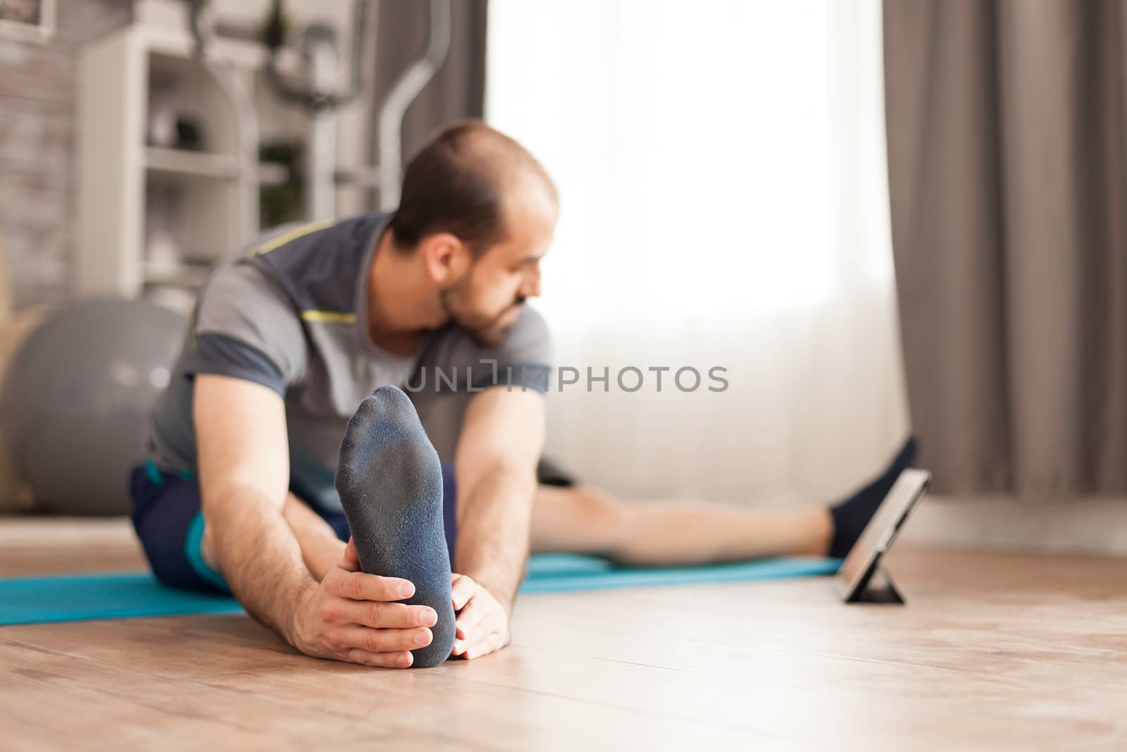 Fit male stretching his body watching online pilates class on tablet computer during global lockdown.