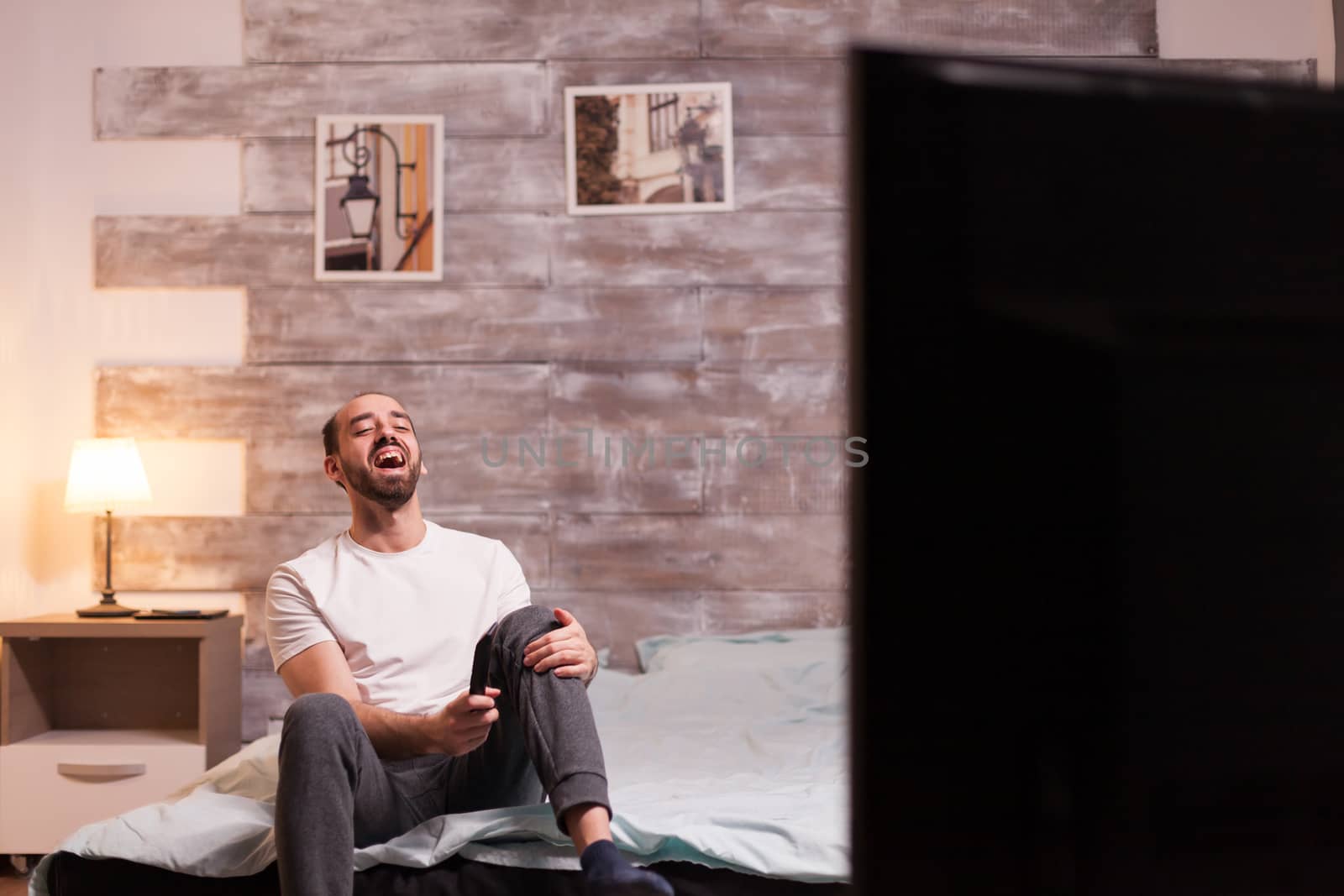 Laughing man on bed at night watching a reality tv show.