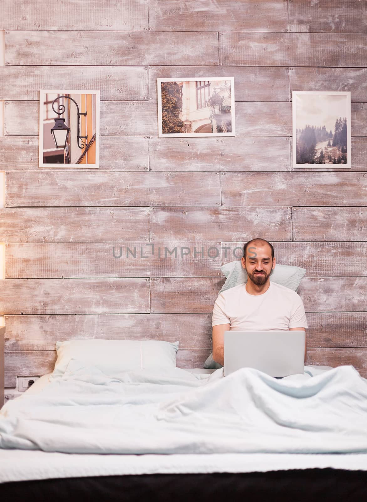 Man smiling while using laptop wearing pajamas in comfortable bed at night.