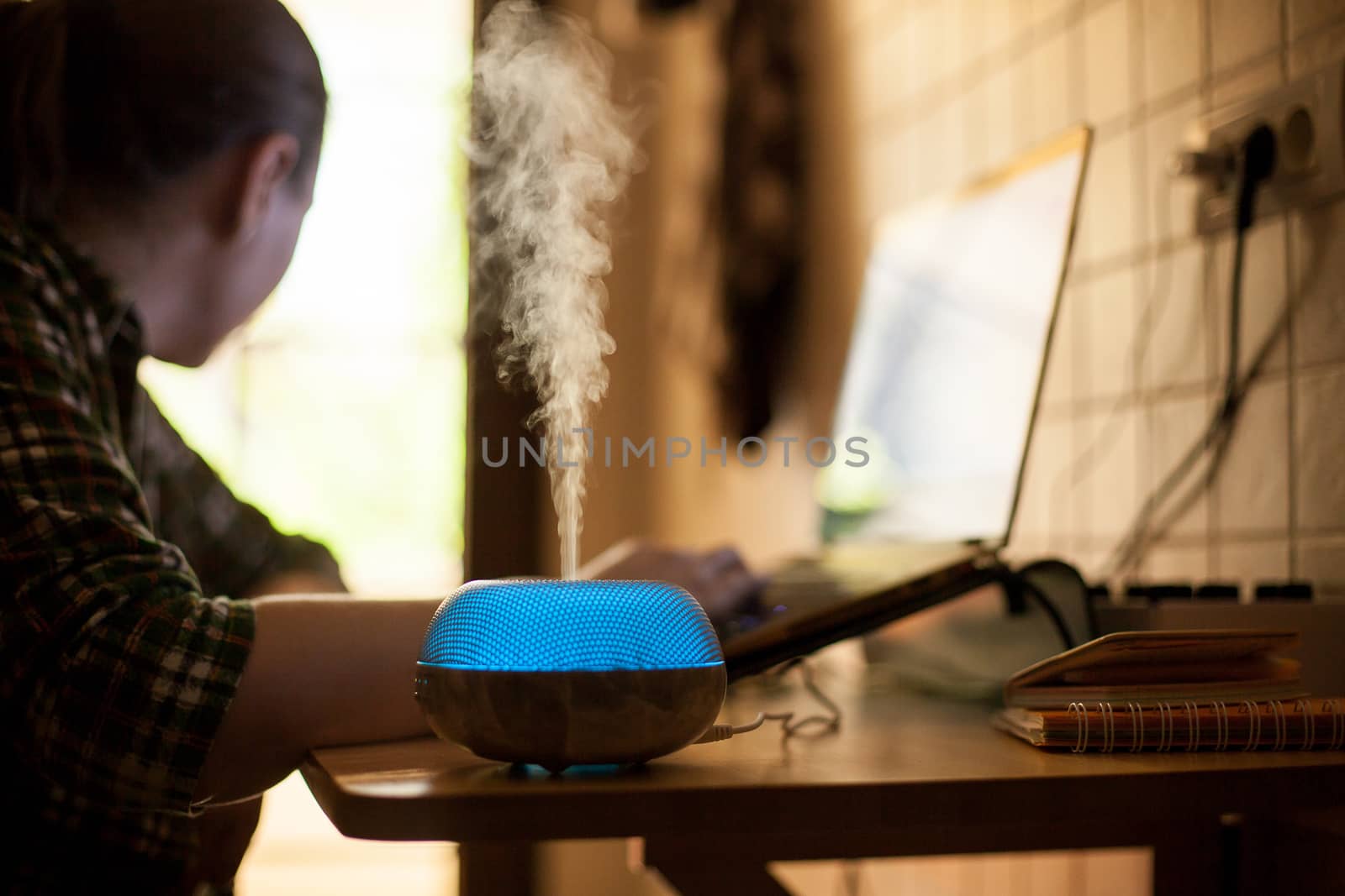 Vapor coming out from essential oil diffureser with blue led while woman working on laptop.