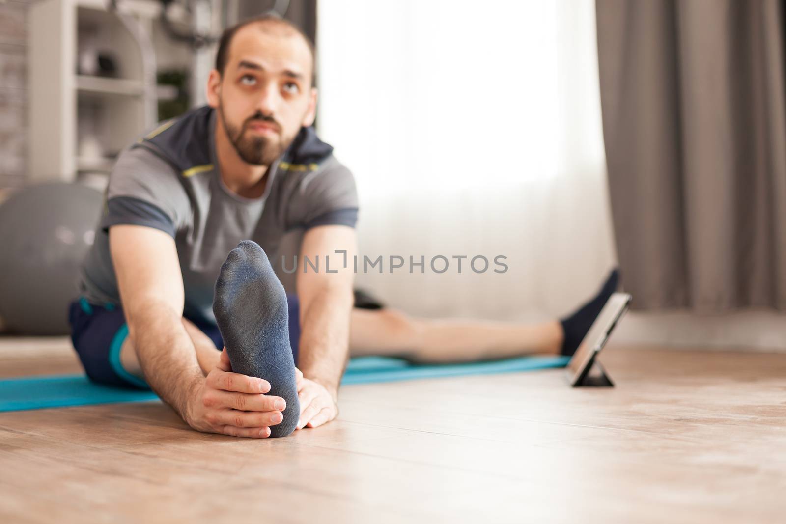 Active man on yoga mat at home stretching his body during online class on tablet computer.