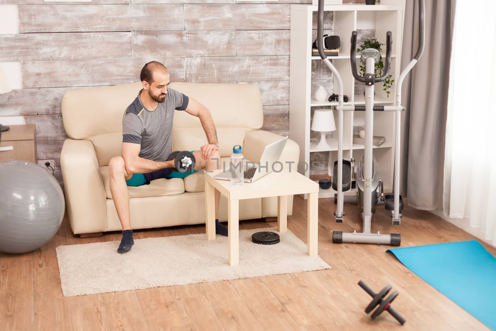 Fit man training his biceps with dumbbell watching online training during global pandemic.