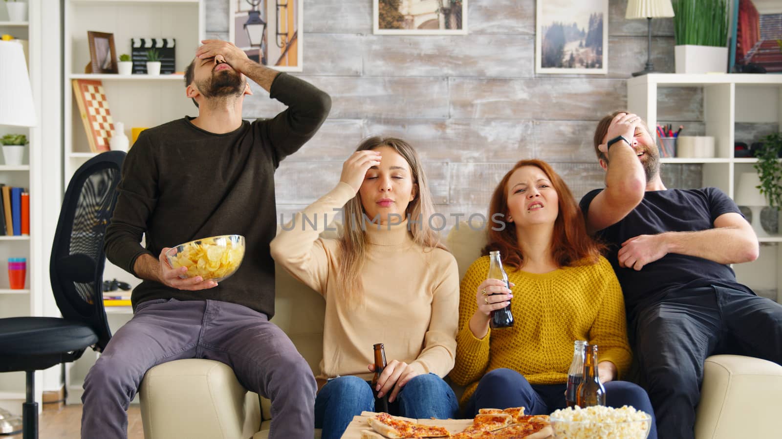 Two couples cheering up while watching a football match on tv in living room drinking beer and eating pizza.