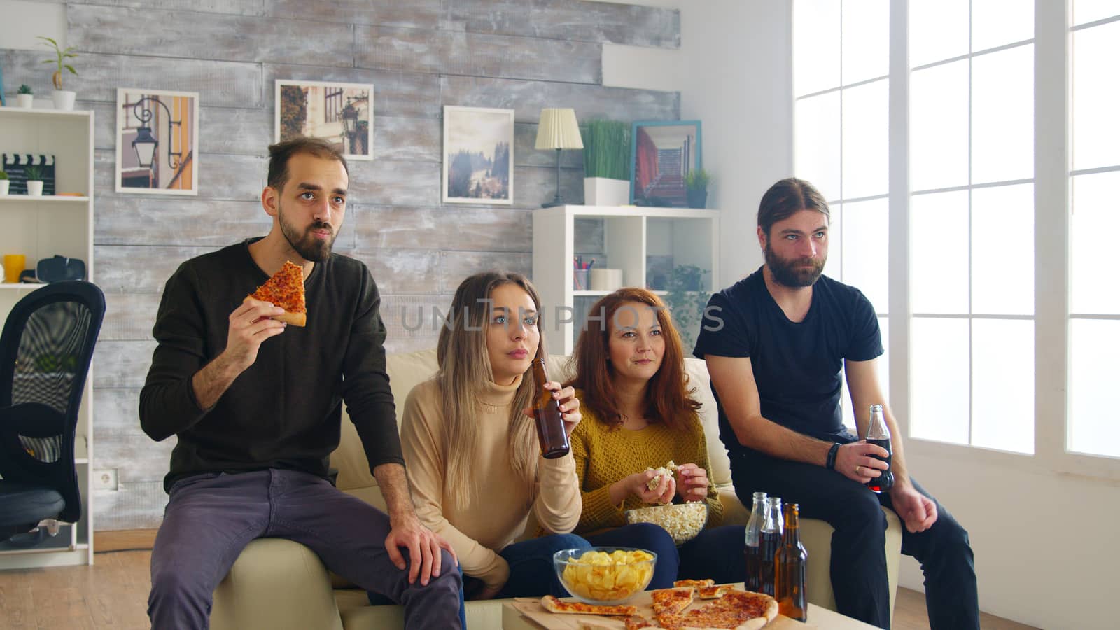 Young man eating chips while playing video games on tv with his friends sitting on the couch. Girl holding a beer bottle.