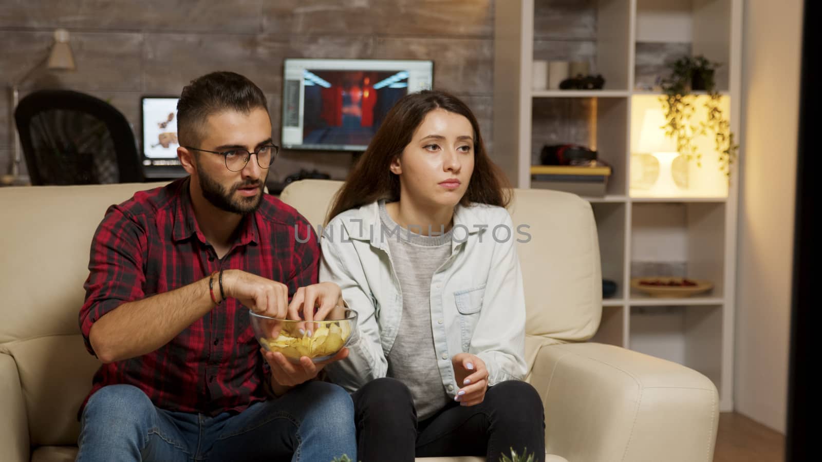Caucasian young couple eating chips while watching television. Couple looking concerned at television.
