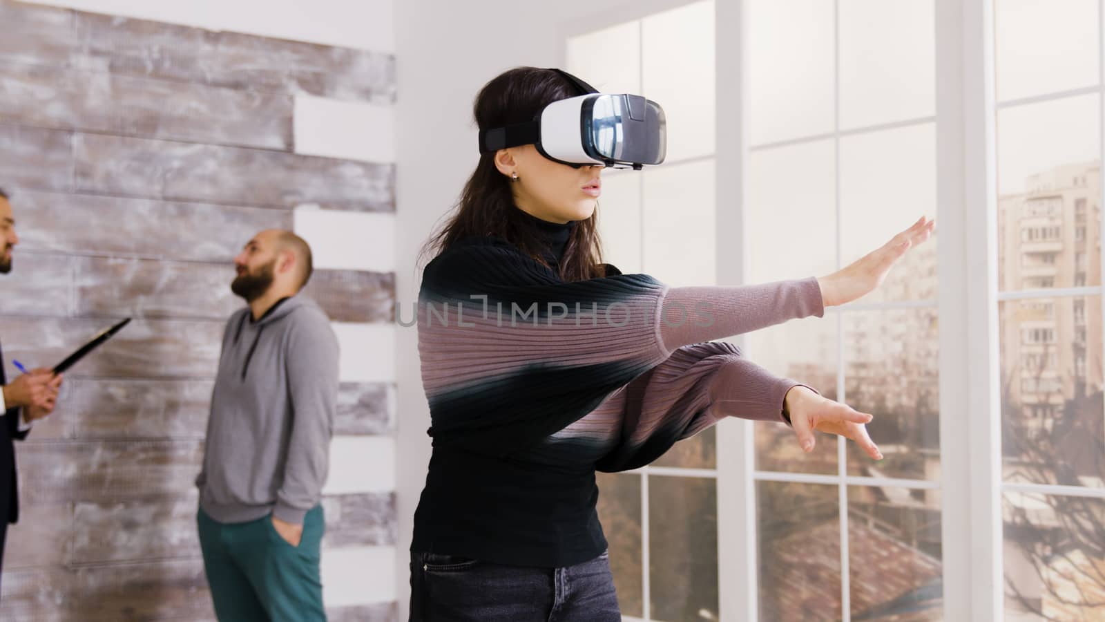 Female architect using virtual reality goggles in empty apartment and real estate agent talking with client in the background.