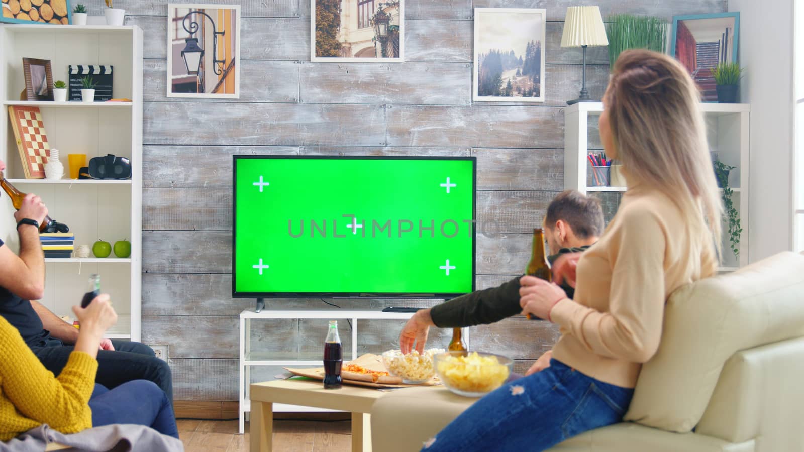 Back view of friends drinking beer and watching sports on tv with green screen in living room.