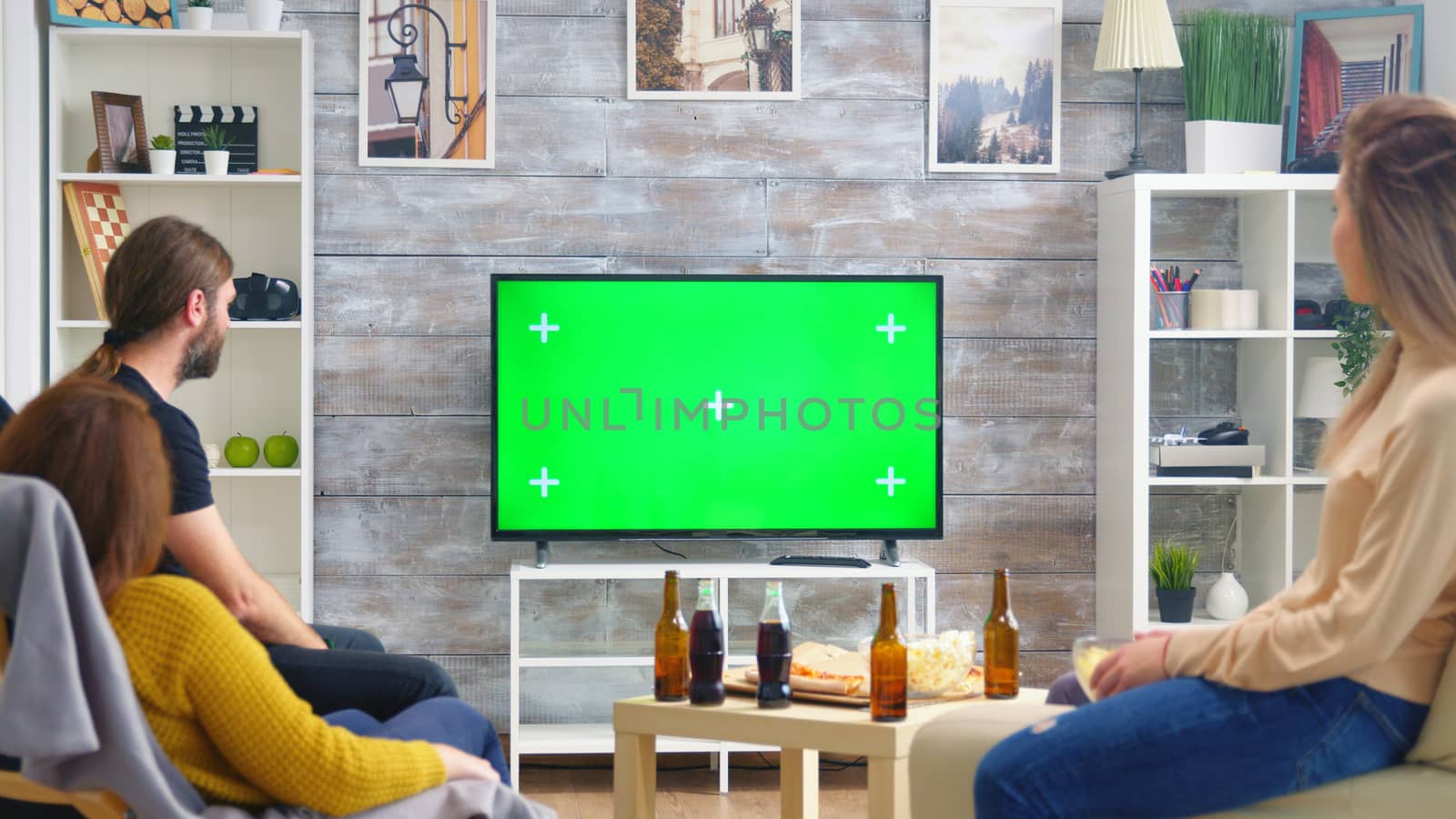 Back view of friends drinking beer and watching sports on tv with green screen in living room.