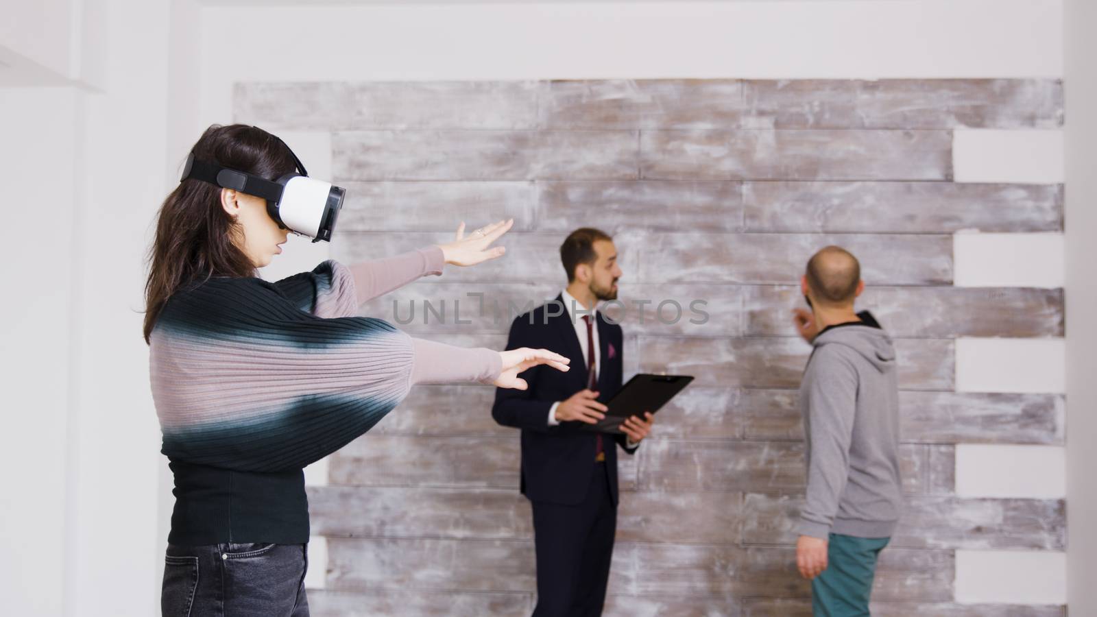 Female designer with virtual reality goggles in empty apartment while real estate agent is talking with the homeowner in the background.