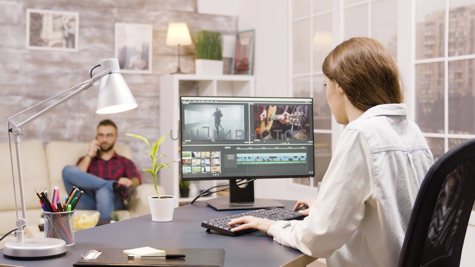 Side view of video editor working on a project from home. Boyfriend in the background is talking on the phone.