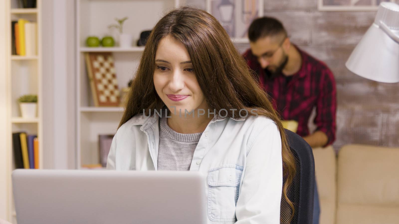 Girl enjoying a cup of coffee while working on laptop in living room. Boyfriend in the background eats chips.