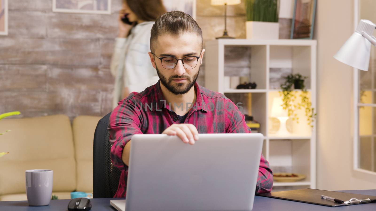 Handsome entrepreneur taking a sip of coffee while working on laptop in living room. Girlfriend in the background talks on the phone.