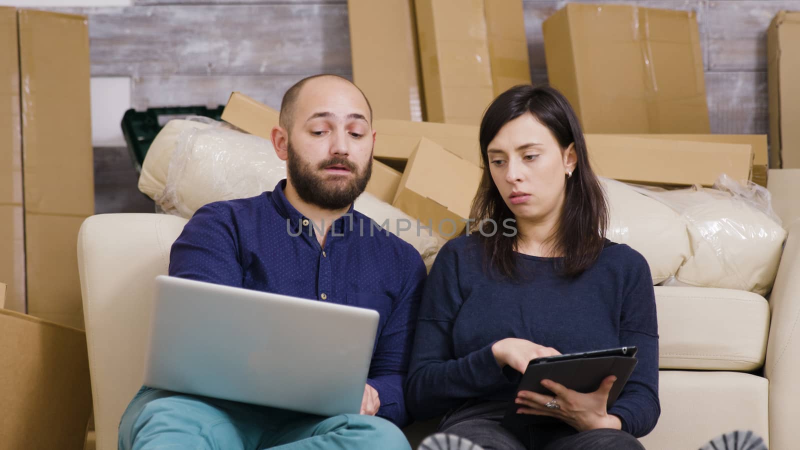 Couple sitting on the floor of their new apartment and making online shopping on laptop and tablet.