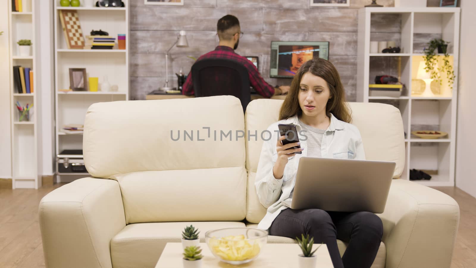 Girl using her phone and laptop while sitting on couch. Boyfriend in the background.