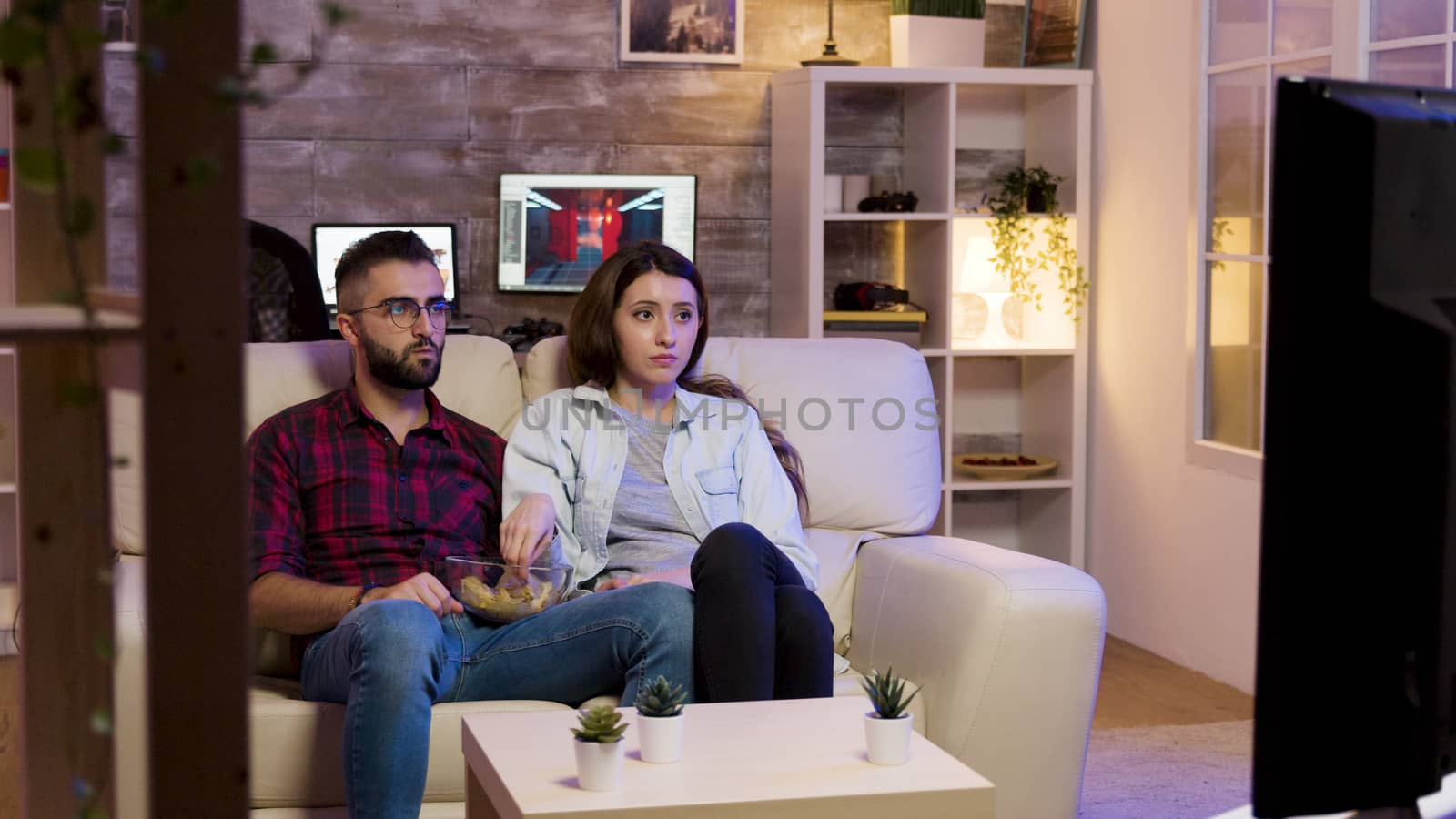 Couple sitting on couch and eating chips while watching a movie on television at night.