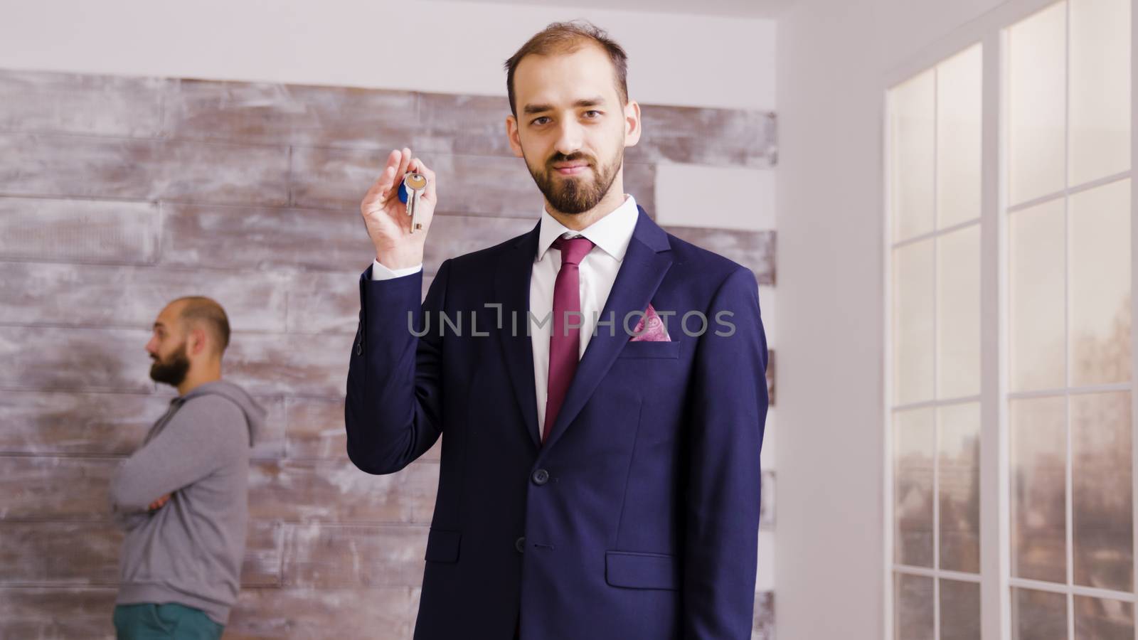 Bearded real estate agent in business suit showing apartment keys to the camera. Couple talking in the background.