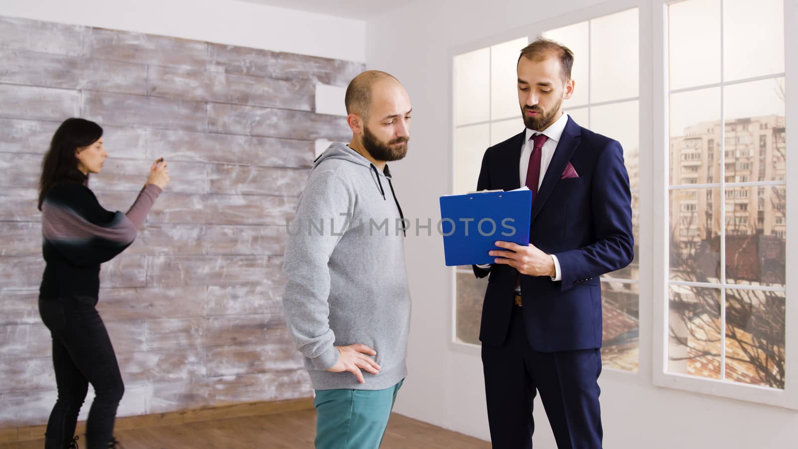 Real estate agent in business suit describes apartment to client in empty property while woman is taking photos in the background.