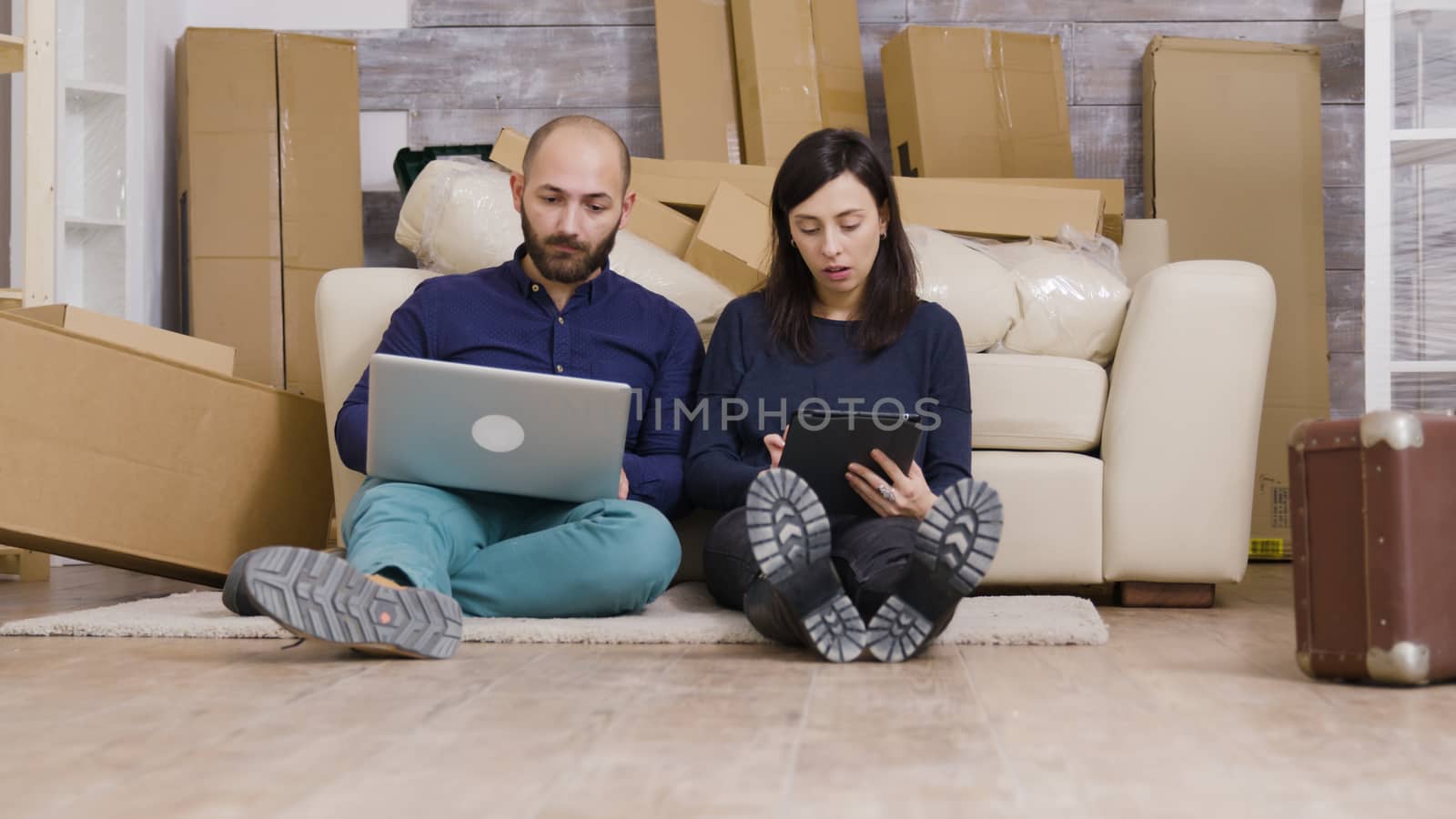 Couple in their new flat sitting on the floor shopping online for furniture using laptop and tablet.