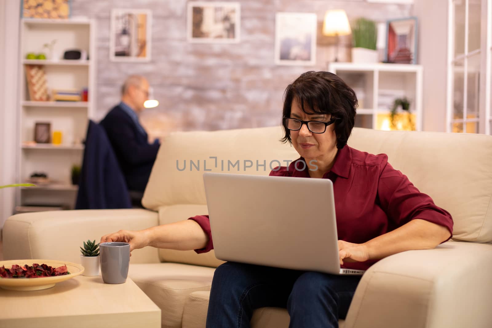 Old elderly woman on her sofa working on a modern laptop in her cozy living room. Her husband is in the background