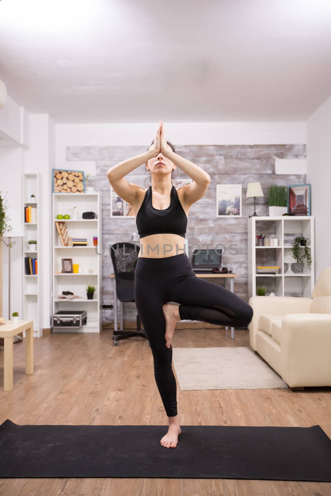 Young caucasian woman practicing tree yoga pose wearing black sportwear in home.