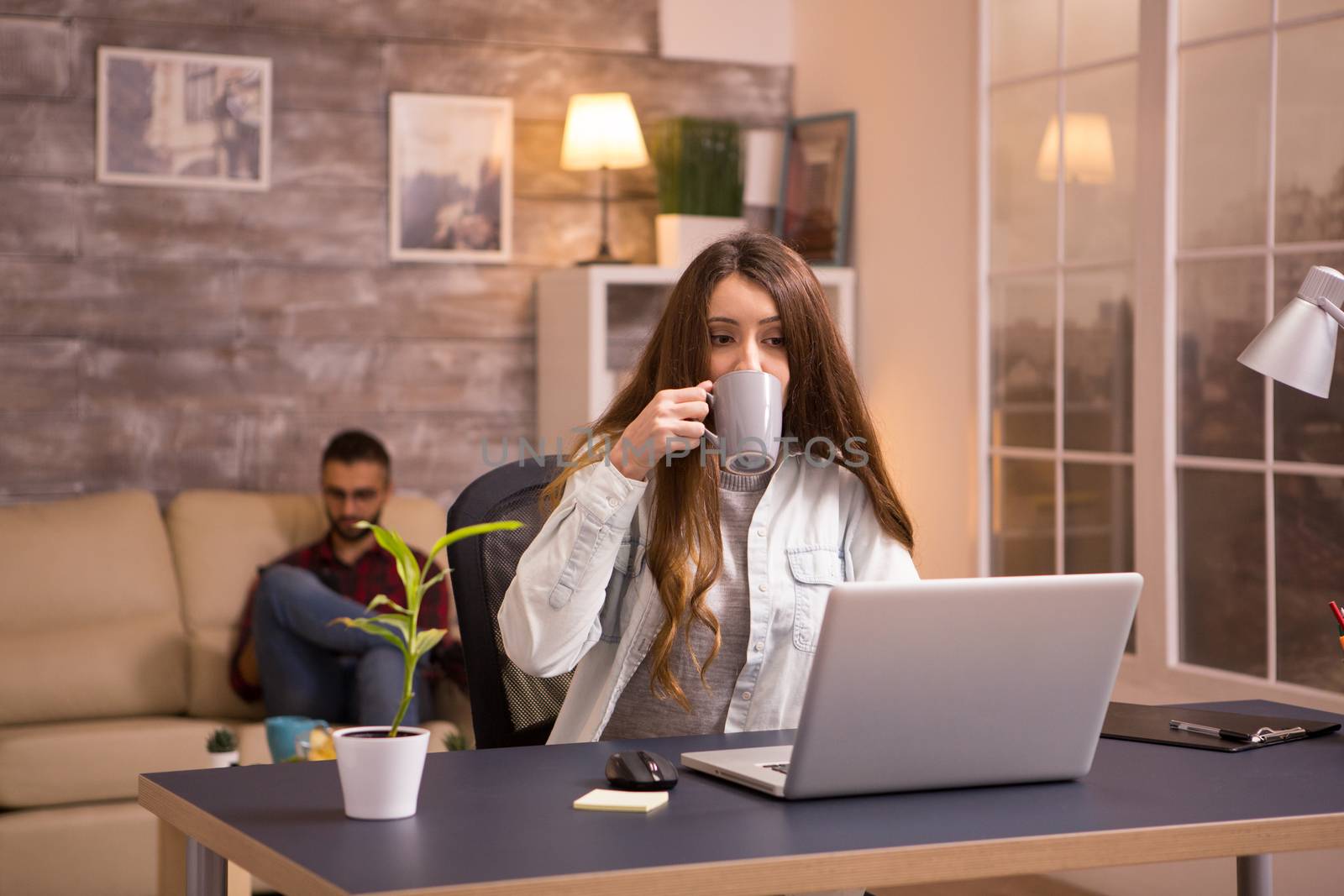 Young female drinking coffee and working on laptop from home office. Boyfriend sitting comfortable on the sofa in the background