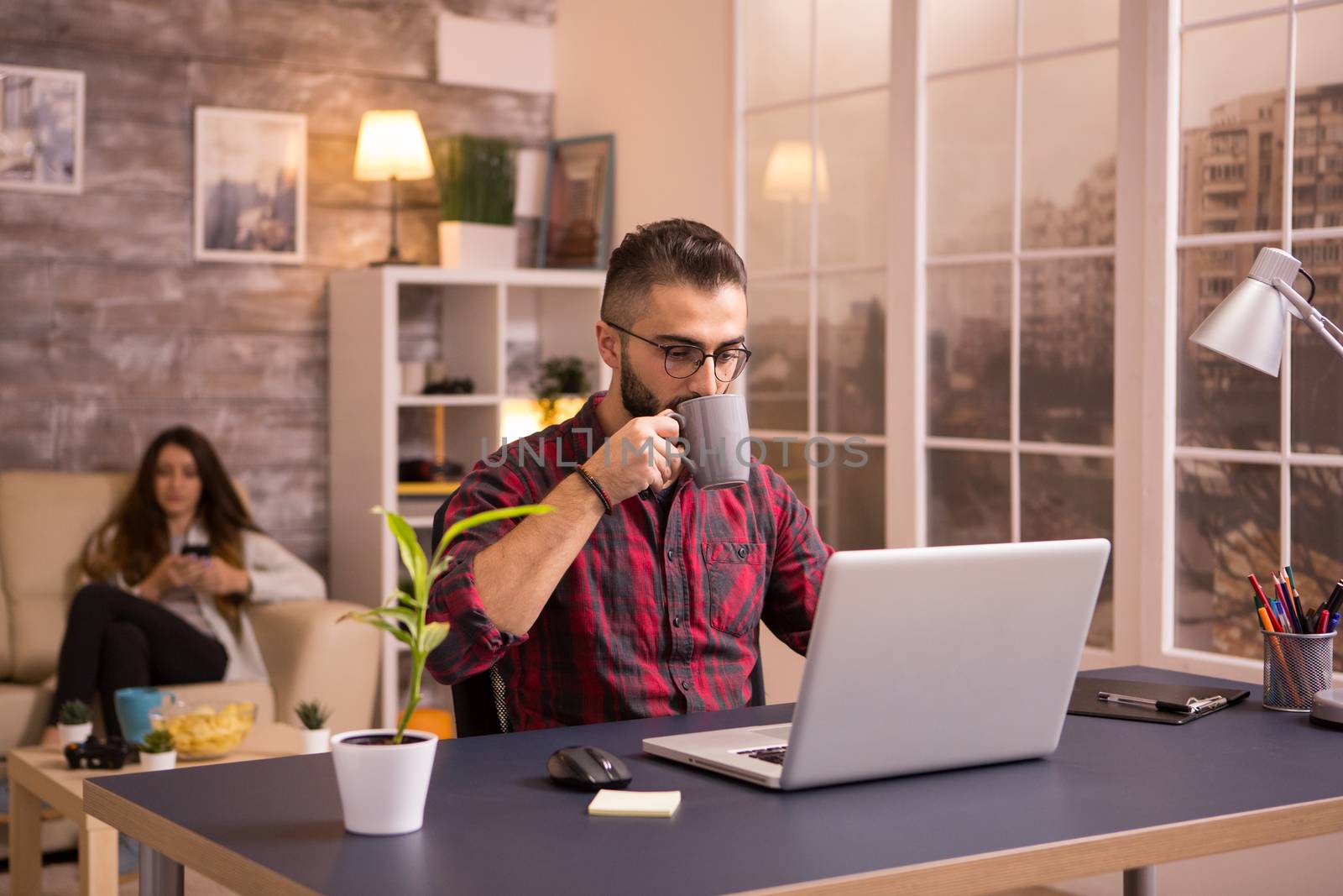 Bearded caucasian entrepreneur taking a sip of coffee while working on laptop in living room. Girlfriend on sofa in the background browsing on phone. Chips on table.