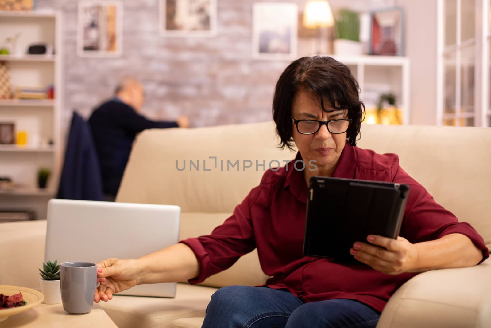 Old elderly woman sitting on the sofa and using a digital tablet PC in cozy living room.