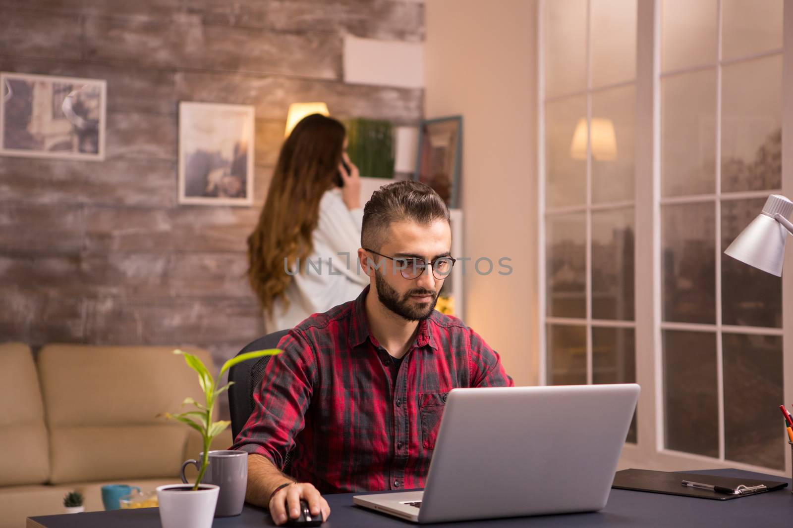 Bearded freelancer working typing on laptop from home. Girlfriend in the background.