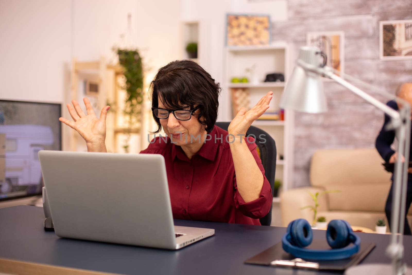 Old woman using a modern computer in her living room while her husband walks in the background