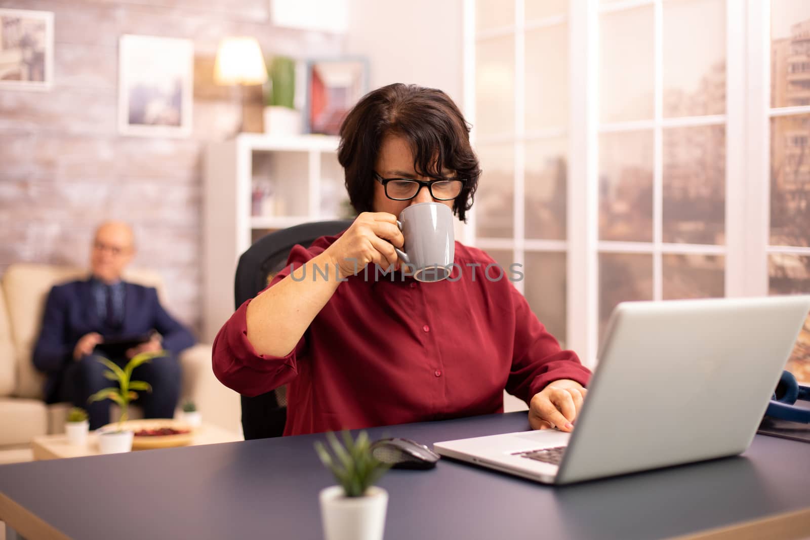 Elderly lady sipss from coffee while working on a modern laptop in cozy living space