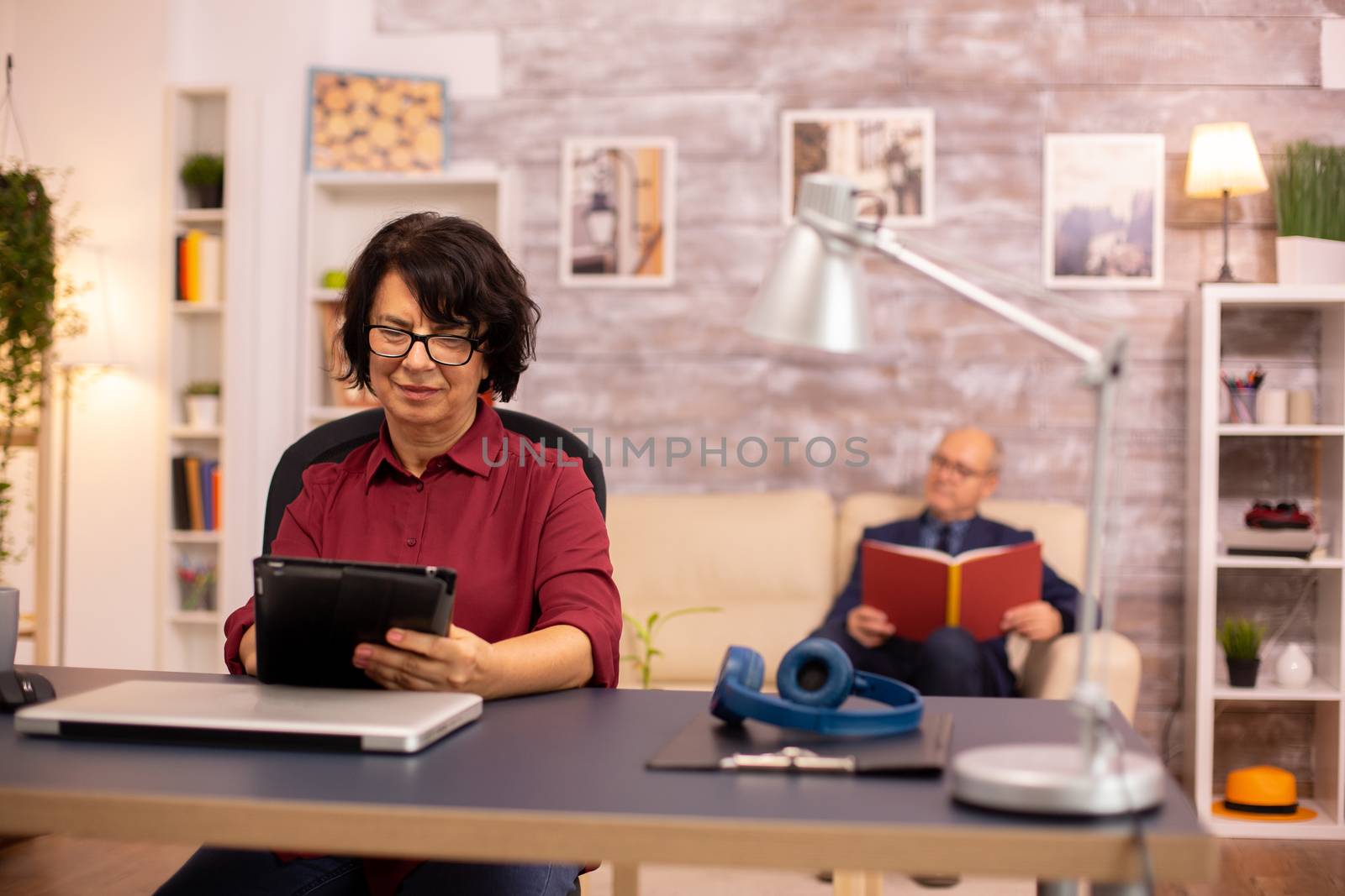 Old lady in her 60s using modern technolgy in cozy living space while her husband reads in the background