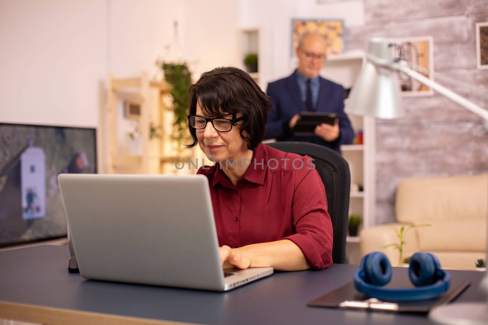 Old woman using a modern computer in her living room while her husband walks in the background