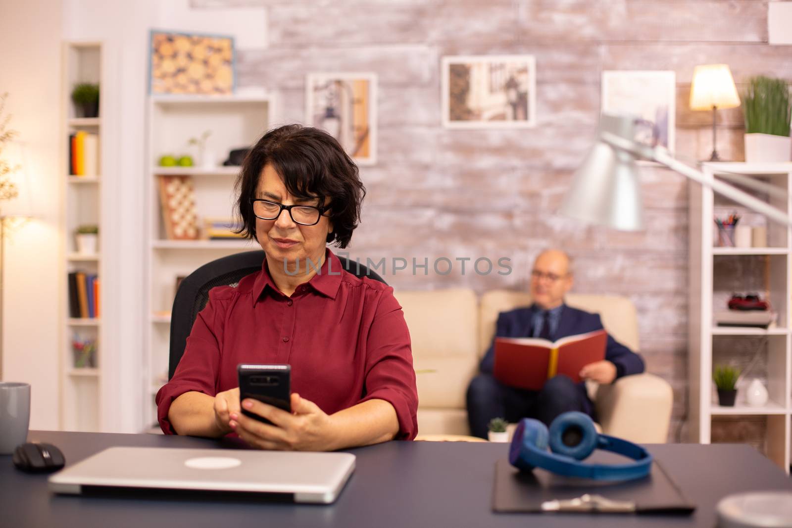 Old lady in her 60s using modern technolgy in cozy living space while her husband reads in the background