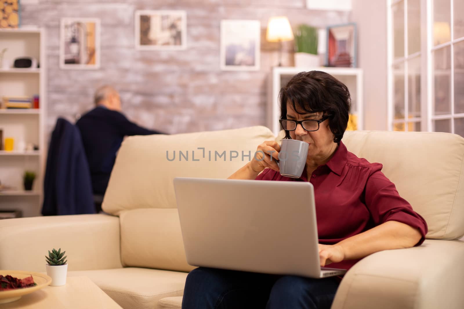 Old elderly woman on her sofa working on a modern laptop in her cozy living room. Her husband is in the background