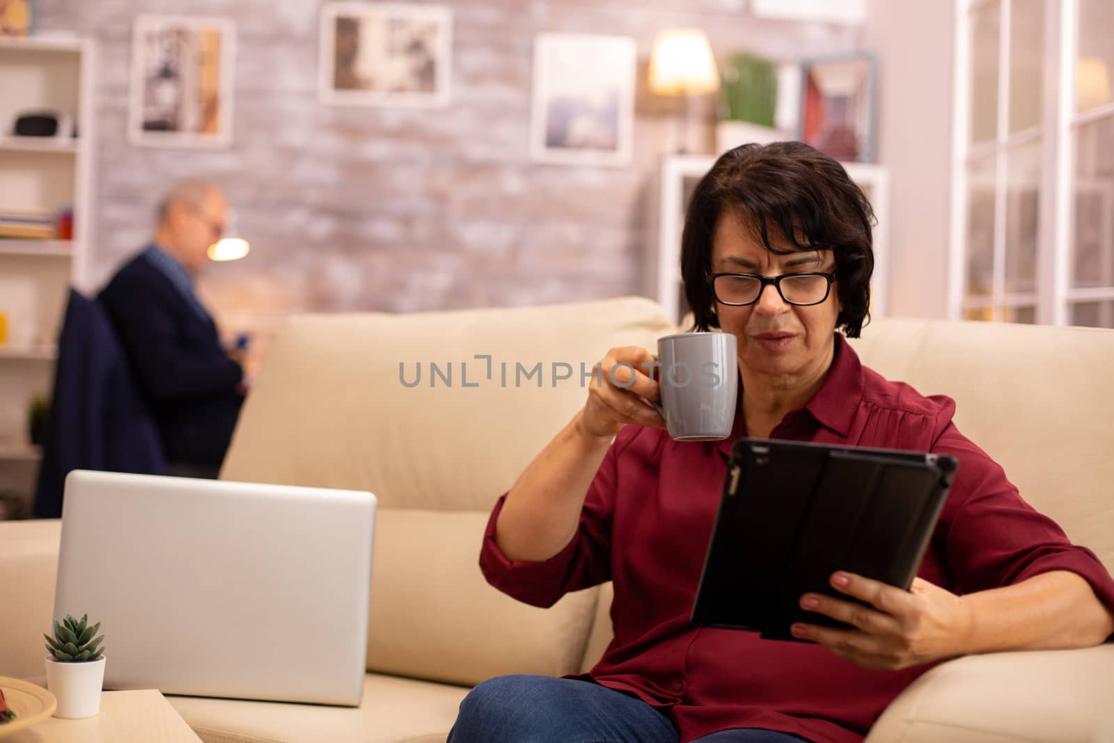 Old elderly woman sitting on the sofa and using a digital tablet PC in cozy living room.