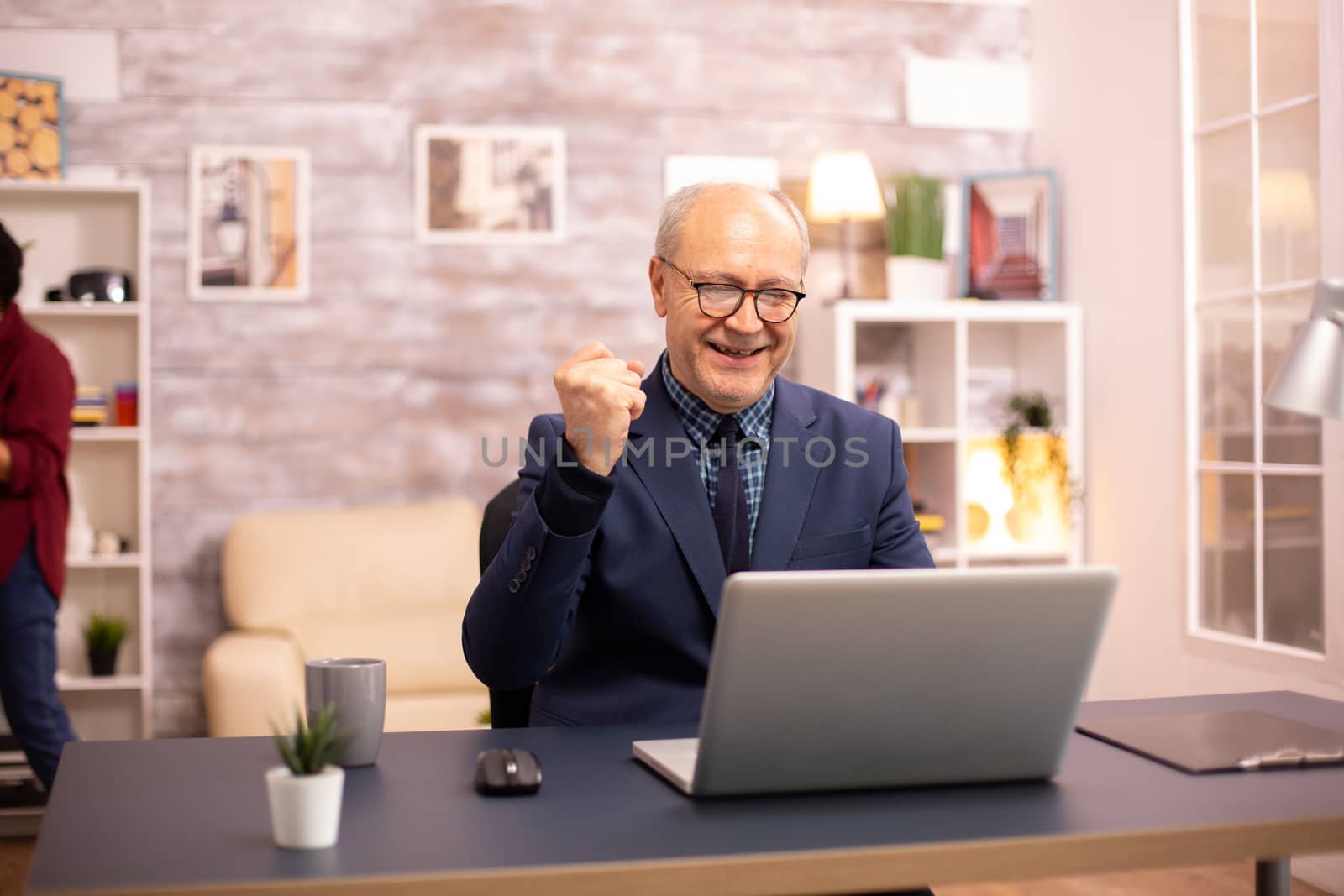 Successful elderly person in suit working on a laptop from home. His wife is in the background