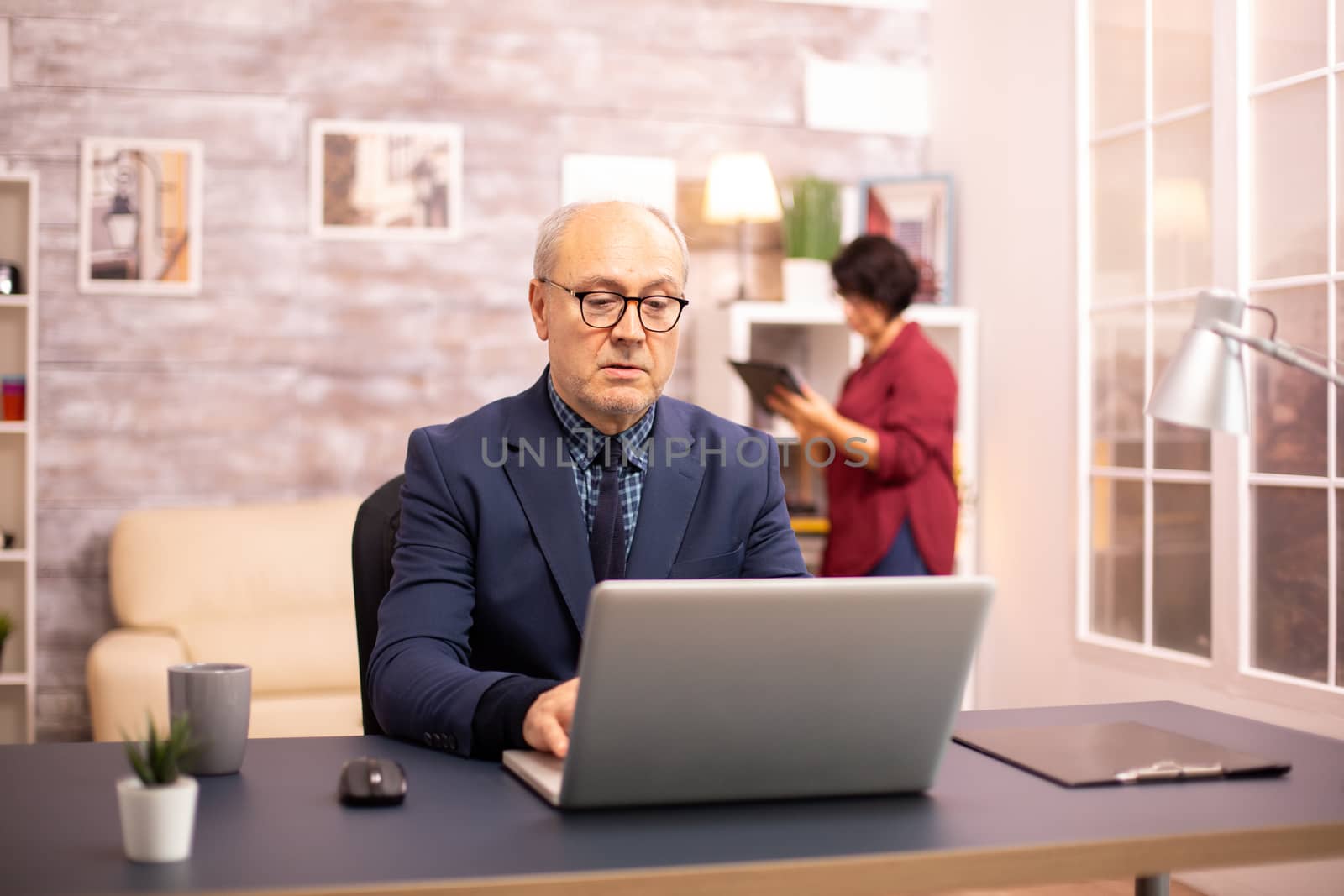 Old man in his 60s working on a laptop in cozy living room while his wife is in background