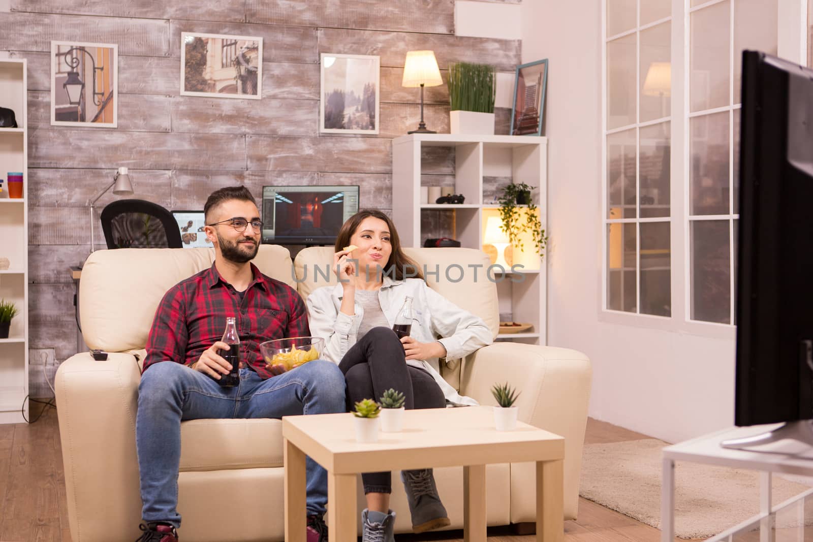 Beautiful young couple drinking soda and eating chips while watching a movie on tv sitting on couch.