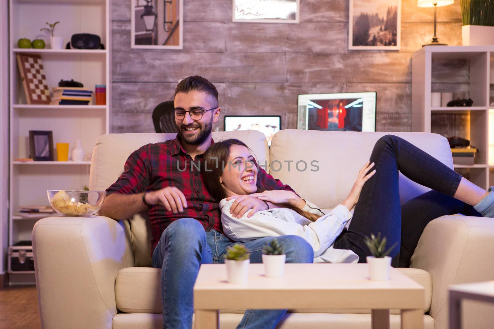 Cheerful young couple laughing while watching a tv show on tv at night. Couple sitting on sofa.