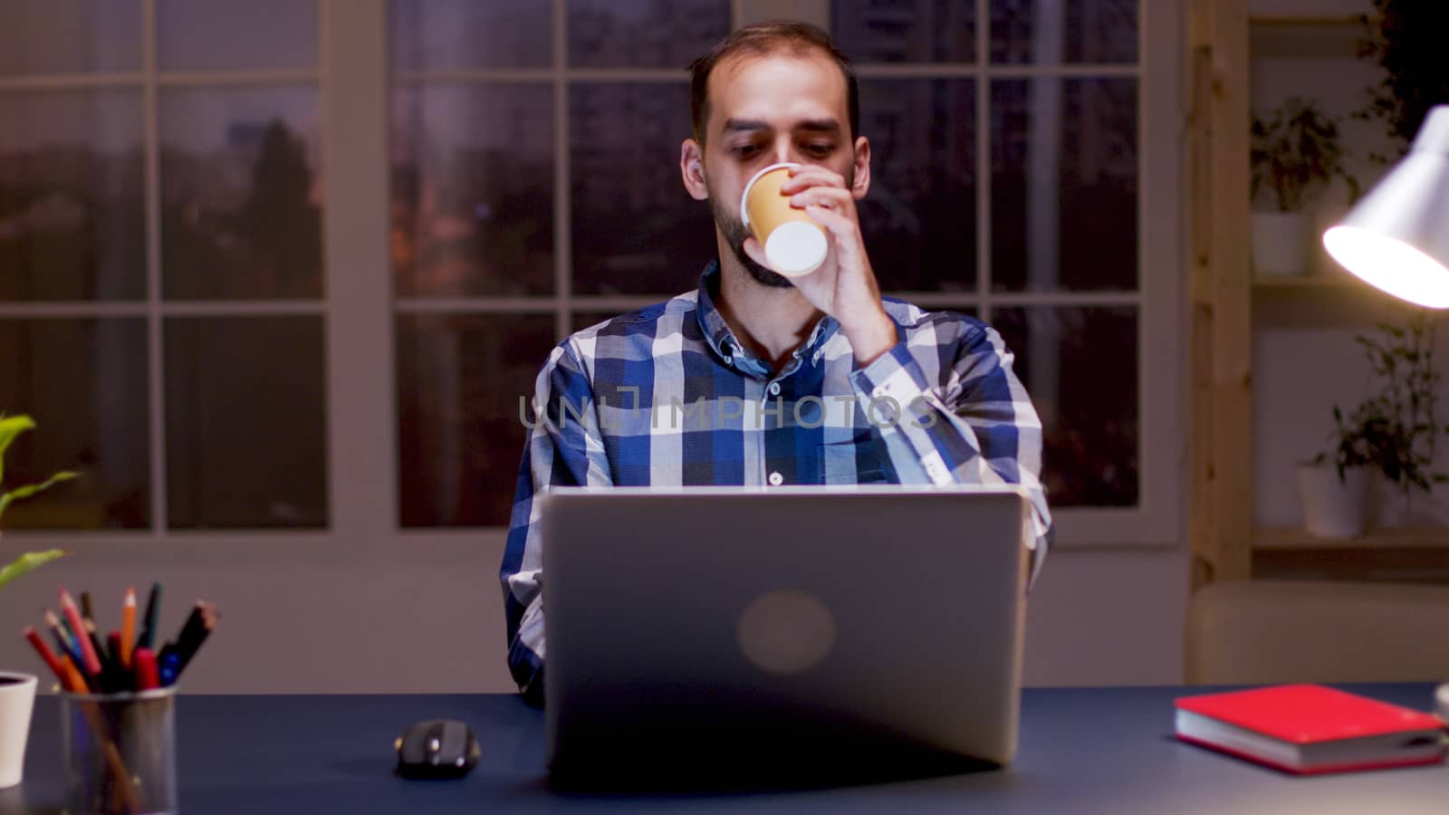 Bearded entrepreneur taking a sip of coffee and working on computer in his home office during night hours.