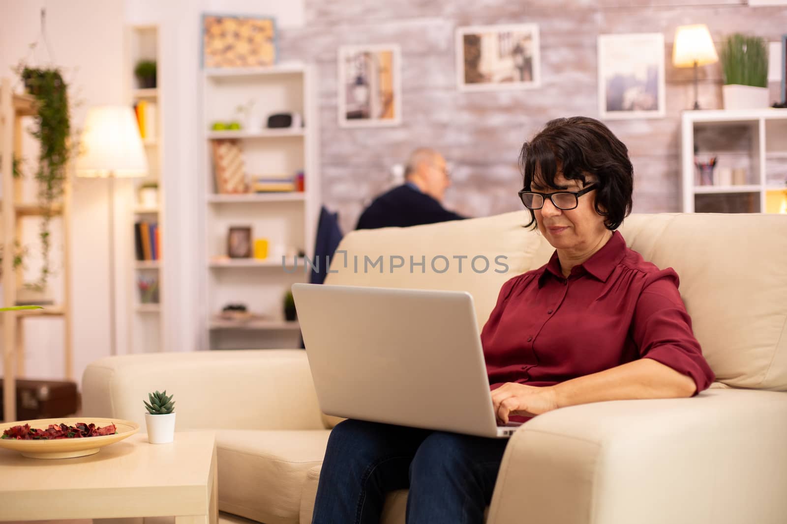 Old elderly woman on her sofa working on a modern laptop in her cozy living room. Her husband is in the background