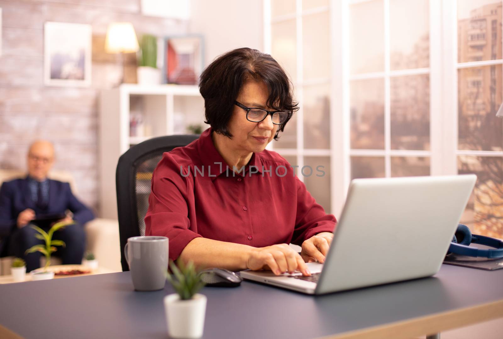 Old woman in her 60s using a modern laptop in her cozy house late in the evening