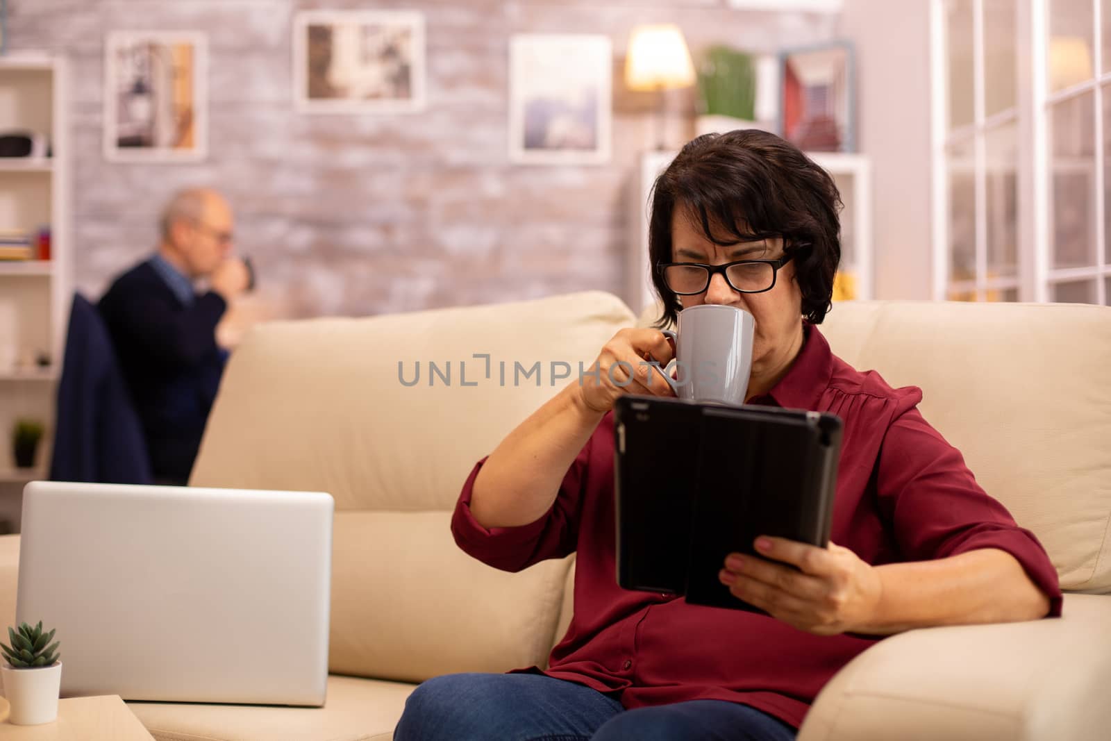 Old elderly woman sitting on the sofa and using a digital tablet PC in cozy living room.