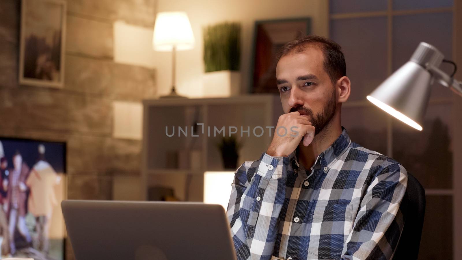 Businessman thinking and holding arms crossed while working on laptop in home office during night hours.