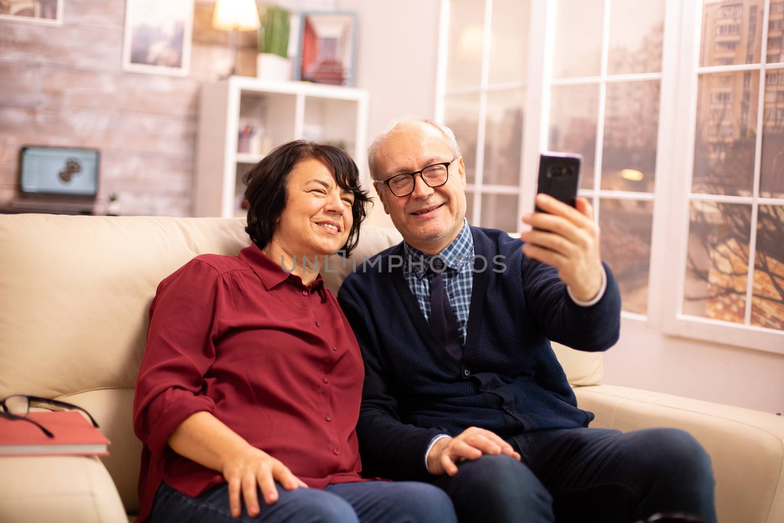 Beautiful elderly couple taking a selfie while sitting on the couch by DCStudio