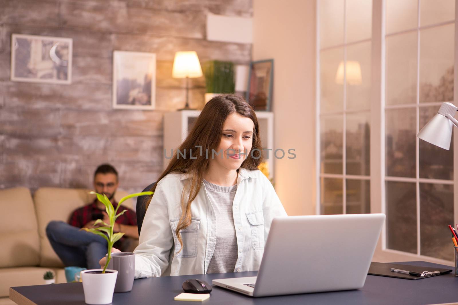 Young woman smiling while working on laptop and holding a cup of coffee. Boyfriend relaxing on sofa in the background.