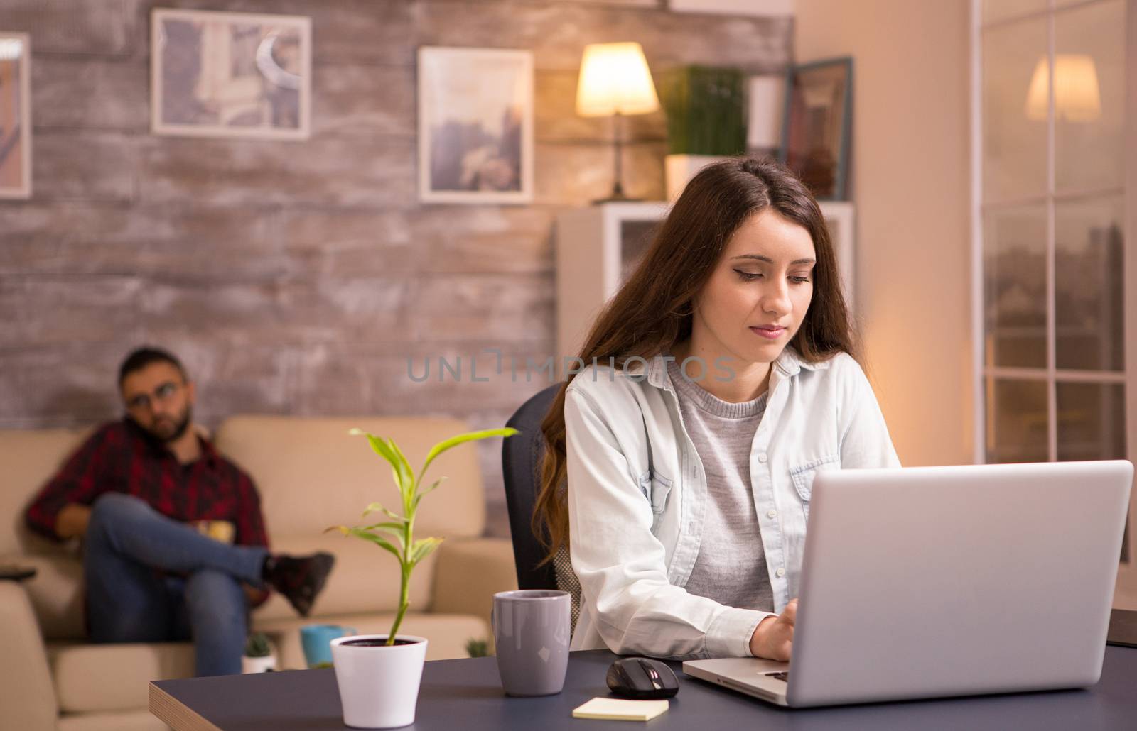 Young female freelance writing and working on laptop in living room. Boyfriend talking on the phone and eating chips in the background.