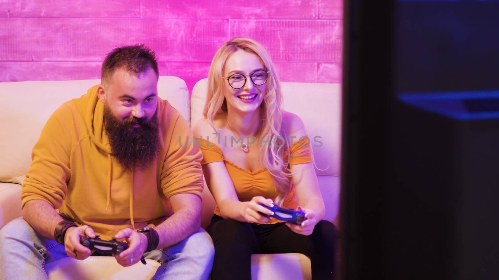 Beautiful blonde girl smiling while playing video games with her boyfriend using wireless controllers on a big screen TV