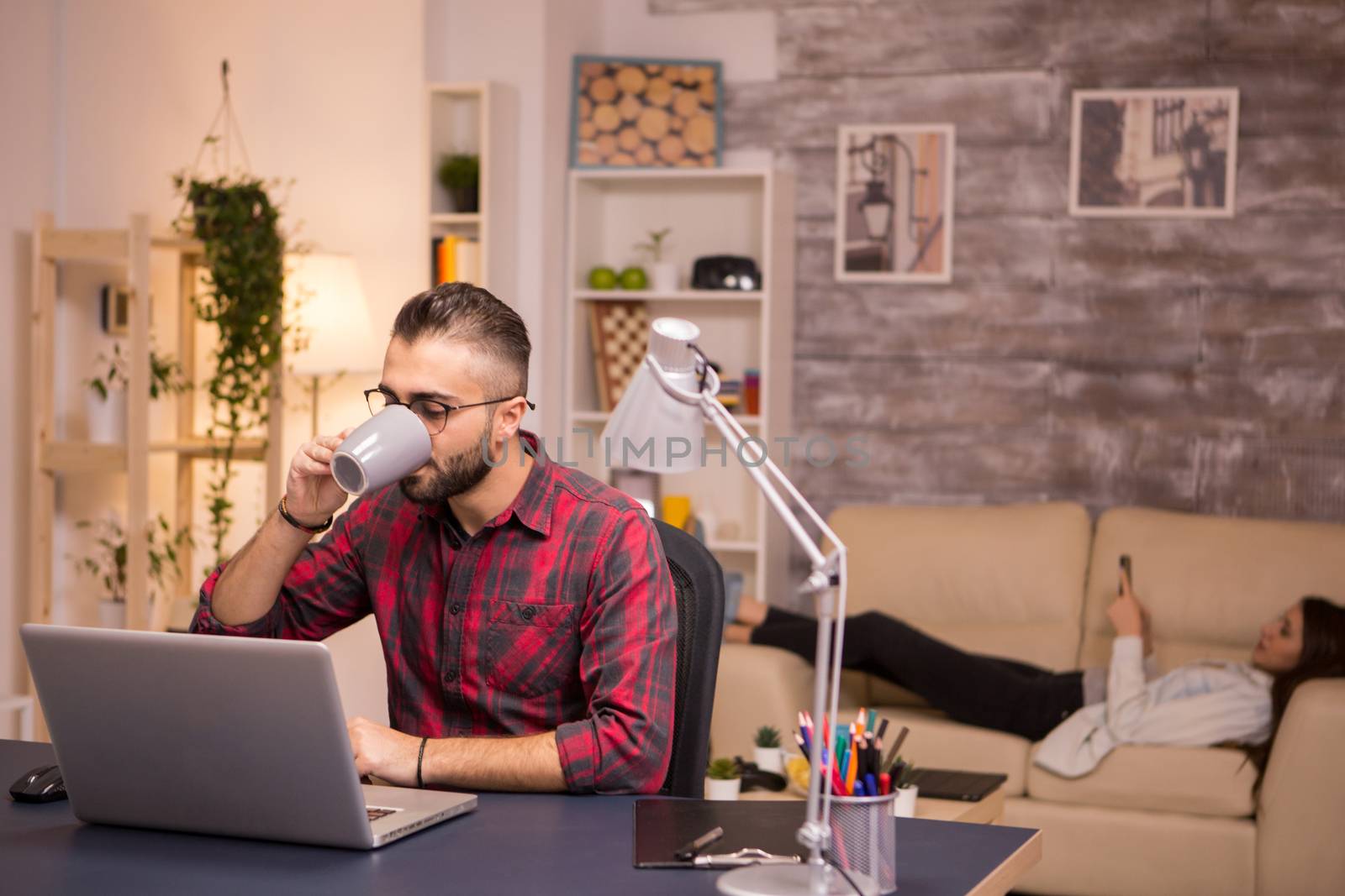 Bearded entrepreneur drinking coffee while working on laptop from home. Girlfriend lying on sofa in the background.